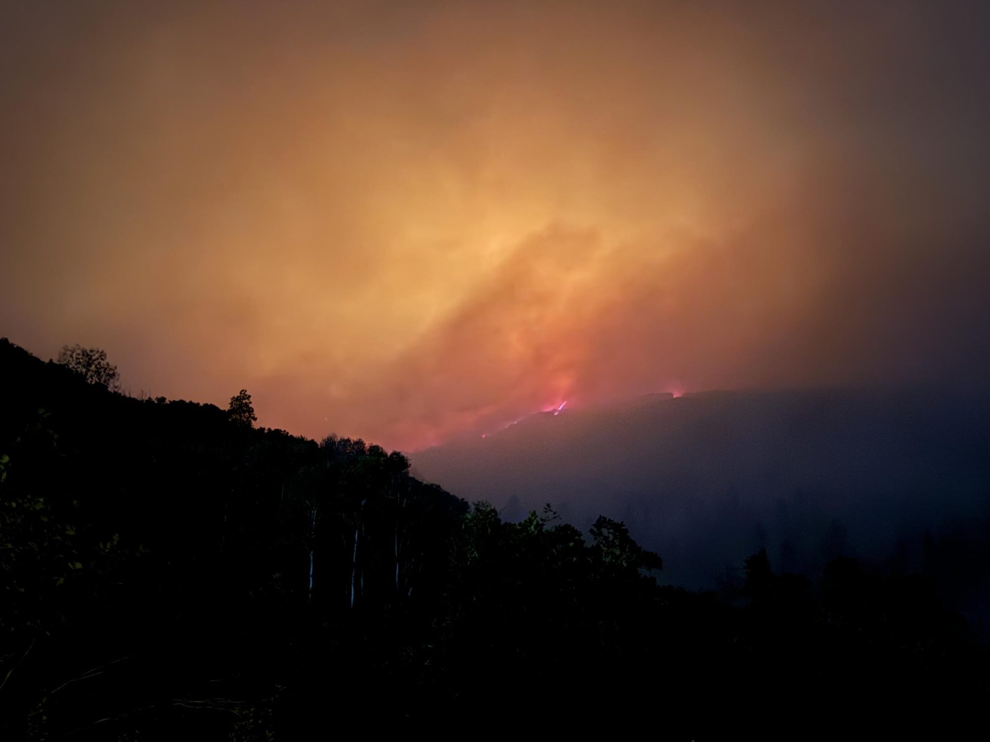 A wildland fire burning along a dark ridge at night, its glow lighting the smoke above the treeline
