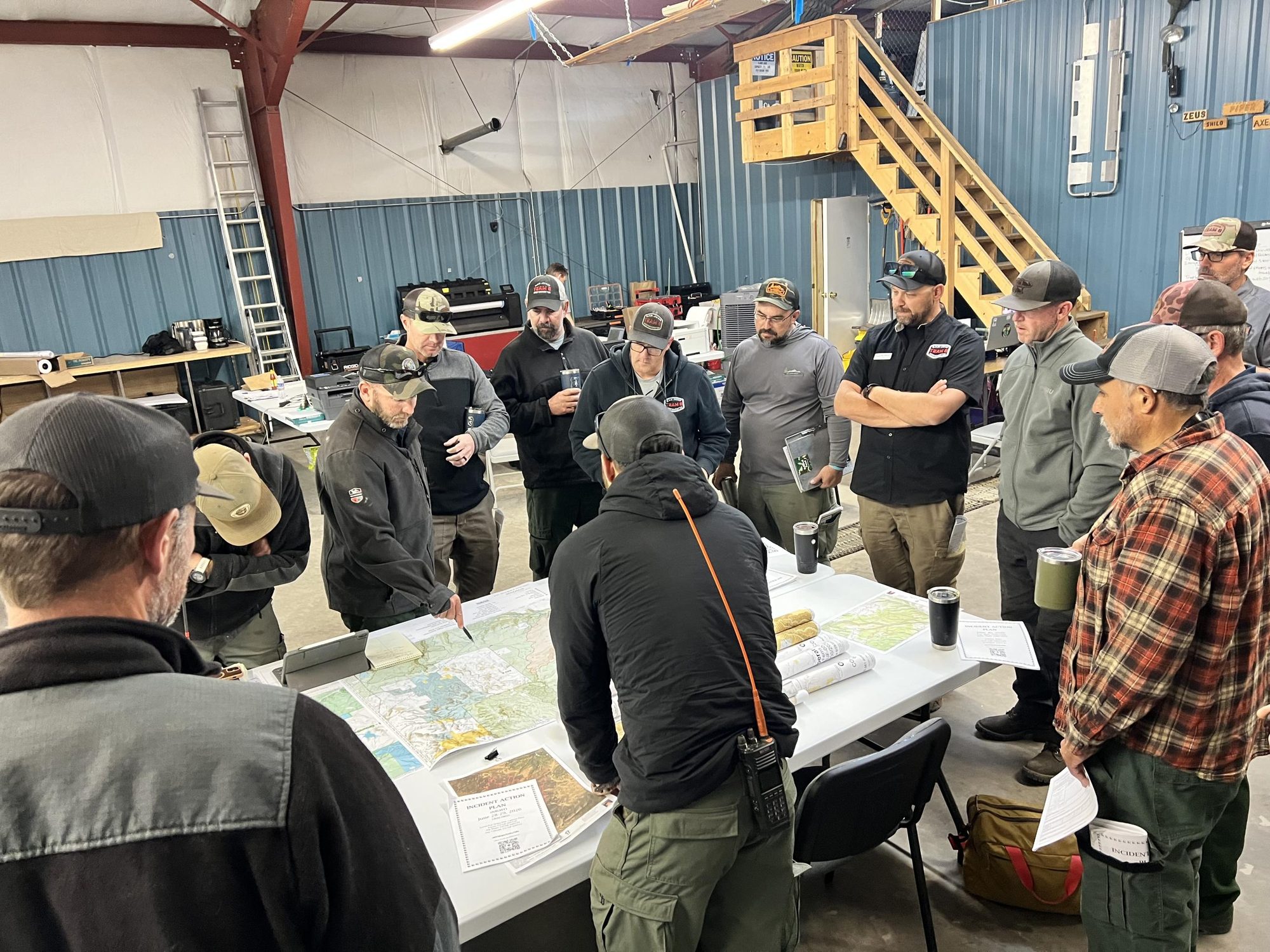 A multi-agency briefing: crews from several organizations standing around folding tables covered with paper maps and a printed incident action plan